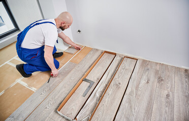 Male builder checking distance from wall to laminate board. Man in work overalls using tape measure while installing laminate flooring in apartment under renovation. Hardwood floor renovation concept.