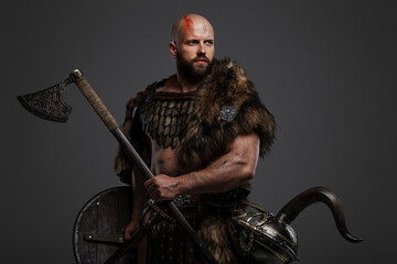 A rugged, bald, bearded Viking dressed in fur and light armor, with a helmet attached to his belt, holding an axe and shield against a gray background