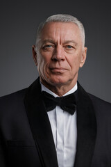 Distinguished older gentleman exudes confidence in this photo, dressed in a sharp black suit and bow tie. The grey background highlights his striking grey hair and determined pose