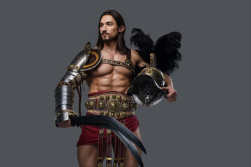 Fototapeta na wymiar Brutal warrior in light armor clenches a gladius and helmet in hands while posing against a gray backdrop