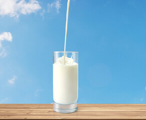 Milk poured into glass on wooden table with sky as background
