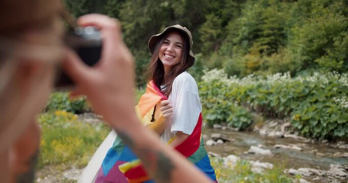 POV shot of a girl taking a picture of her friend who is standing with an LGBT flag in a white T-shirt against the backdrop of a green forest