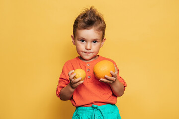 Studio shot of a smiling boy holding a fresh lemon and orange on a yellow background. The concept of healthy baby food, vitamin C.