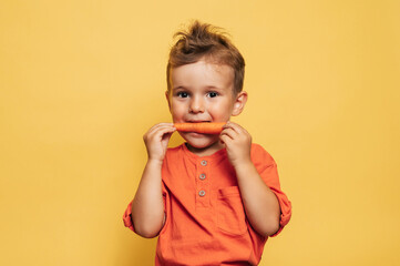 Studio shot of a smiling boy eating fresh carrots on a yellow background. The concept of healthy...