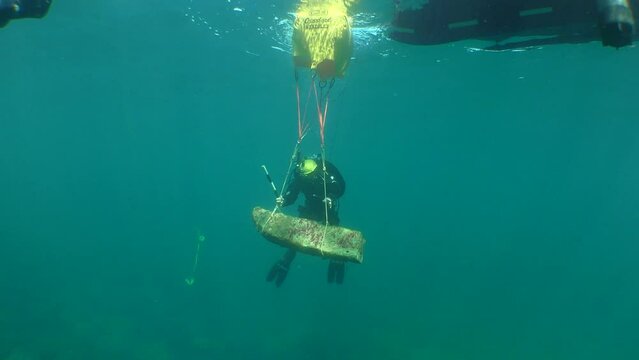 Underwater archeology: a bag of an underwater lifting system attached to an antique Greek stone anchor on the surface of the sea, near a diver, wide shot.