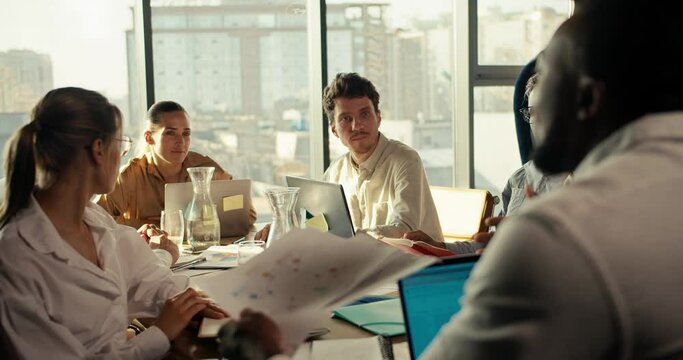 An office worker with black skin color in a white shirt tells his business plan to colleagues at the office table in a room with large windows