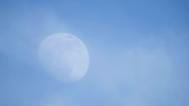 The massive moon graces daylight skies, drifting through clouds with an enigmatic haze.
