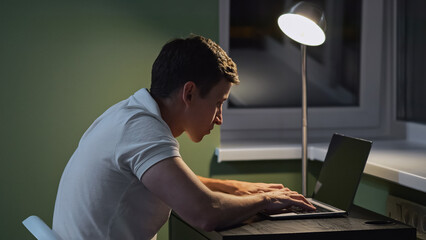 Male person with incorrect posture types on laptop keyboard near floor lamp. Crooked posture and health problems. Remote work at home during night - obrazy, fototapety, plakaty