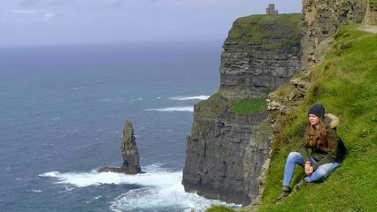 Young woman siitting at the Cliffs of Moher at the Irish west coast - slow motion clip  - Powered by Adobe