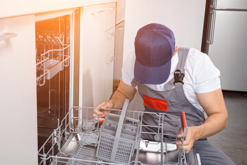 Male plumber in uniform inspects dishwasher part to repair in modern kitchen. Concept of broken...