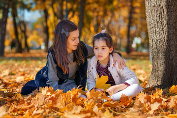 two girls are sitting in a glade of yellow maple leaves in an autumn city park, children are playing and enjoying, picking leaves near a tree, beautiful nature, bright sunny day