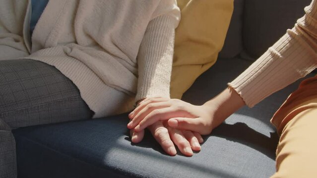 Cropped shot of unrecognizable woman holding hand of senior grandmother while sitting together on couch during day at home