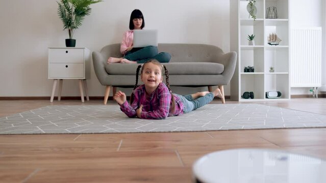 Preteen girl switching on robot vacuum while resting on carpet with occupied lady in background. Smart daughter starting daily hoovering with modern technologies while mom doing tasks for employer.