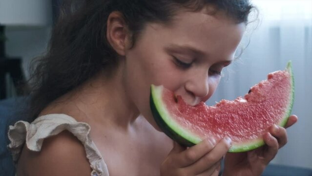Close-up of preteen girl eating watermelon fruit sitting on comfortable couch at home. Happy cute kid snacking fruit. Childhood and healthy food concept. Pretty little child eats healthy snack food