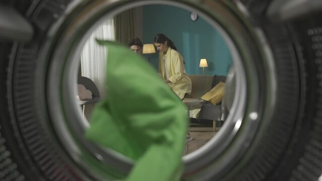 View from inside the empty washing machine young couple with laundry basket throwing clothes inside. Slow motion HDR BT2020 HLG Material.