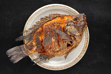 tasty large fried nile tilapia fish in circle ceramic plate on dark tone texture background, top view, flat lay