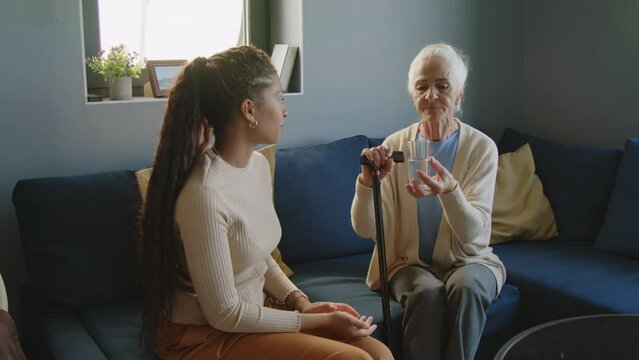 Senior woman sitting with walking cane at home, taking pills and drinking water under control of young female caregiver
