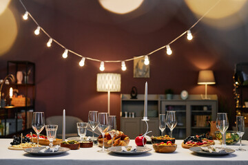 Dinner table served with appetizing homemade food, candles, empty wineglasses and flutes standing in front of camera in living room