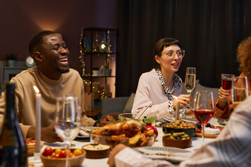 Happy young African American man laughing while sitting by served table next to his girlfriend with flute of champagne during dinner