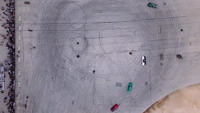 Overhead Drone View of Three Modified Performance Automotive Fast Furious Drift Cars Burning Out Race Event Burning Rubber on Asphalt Parking Lot Track Video Opener with Bystanders