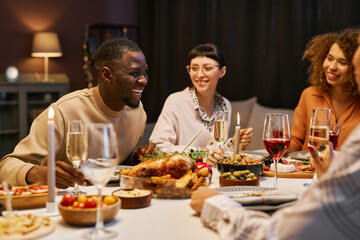 Laughing black man with flute of champagne bending over served table during chat with his friends while enjoying home party and dinner