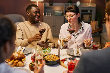 Laughing guy with glass of red wine looking at his girlfriend making toast while both sitting by served table in front of friends at party