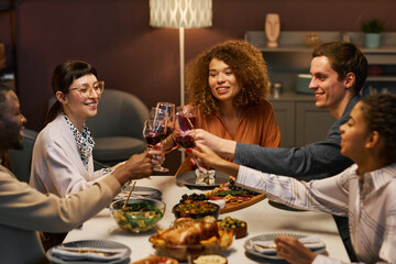 Group of young happy friends clinking with glasses of alcoholic drinks over served table while sitting in front of each other during festive dinner
