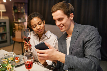 Young smiling man showing curious video in smartphone to his girlfriend with glass of red wine sitting next to him by served table during dinner