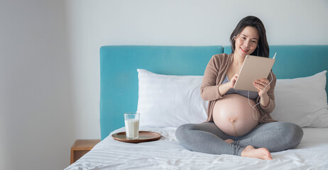 The Asian pregnant woman is listening to music and using her phone while on the bed in the bedroom.