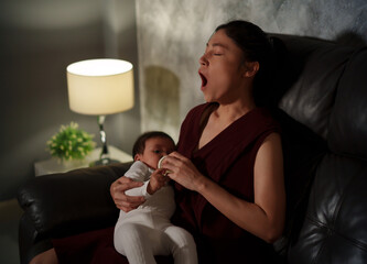 sleepy mother yawning and feeding milk bottle to infant baby on sofa in the living room at night