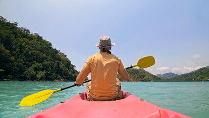 Young man with sunglasses and hat rows pink plastic canoe along sea against green hilly islands...