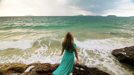 Seductive long-haired woman walks on rock of sea reef stone, stormy cloudy ocean. Woman in blue swimsuit dress tunic. Concept rest in sea, tropical resort coastline traveling tourism summer holidays