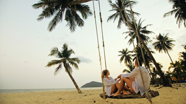 Cute loving couple on the swing on tropical beach. Woman and man in white shirt enjoy gold sunset and life in beaches, palm tree. Evening warm sunset. Summer holiday vacation tropical tourism concept
