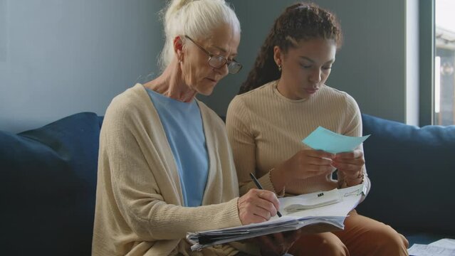 Young female caregiver reading bills and talking to elderly woman making notes in personal financial workbook while planning budget together at home