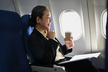Successful middle aged businesswoman drinking coffee and looking through window of the airplane during flight