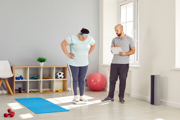 Male personal trainer checking progress of overweight woman in gym. Plus size woman in sportswear weighing on scale in fitness center. Healthy lifestyle, slimming and dieting concept