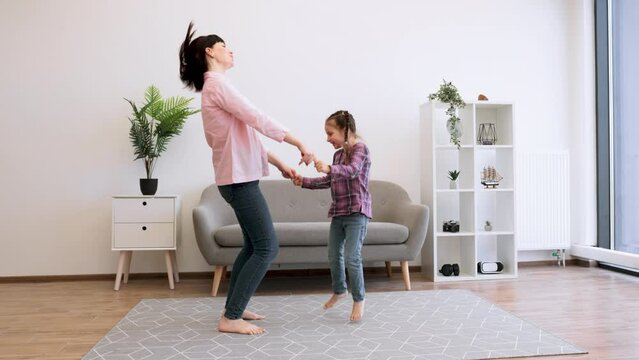 Attractive young lady in casual wear holding arms of cute tween girl while dancing barefoot on carpet in lounge. Cheerful mother and daughter learning new steps while listening to favourite songs.