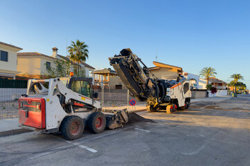 Obraz premium Road construction and asphalt repair in Spain. Bobcat skid-steer loader on roadwork in town. Cold milling machine on asphalt removal and concrete pavement. Road Machinery for Road construction..