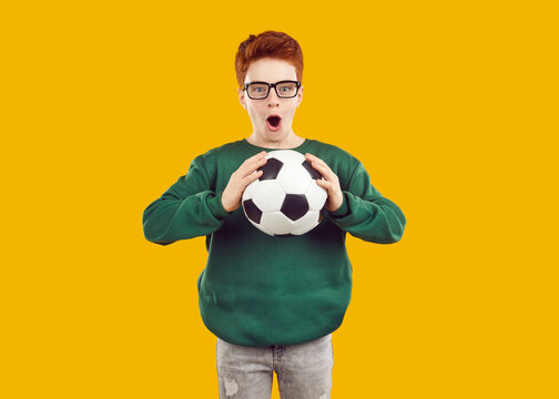 Portrait of excited young boy in glasses holding soccer ball with shocked face and open mouth on yellow background. Studio photo of football fan guy looking to camera with surprised face expression.
