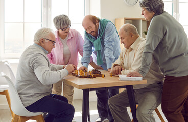 Group of happy diverse senior people playing chess in nursing home sitting at the table. Pensioners spending leisure time together playing board games. Leisure in retirement home concept.