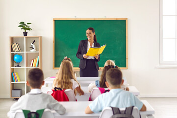 Friendly smiling school teacher with book on lesson in elementary school with her children...