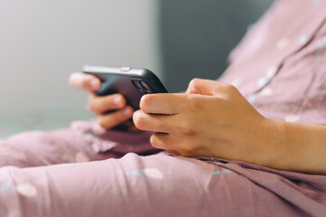 Close up woman hand in pajamas lies on a bed looking at a smartphone screen. Streaming watching leisure concept