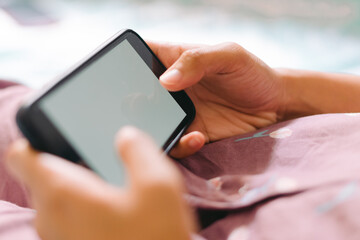 Close up woman hand in pajamas lies on a bed looking at a smartphone with a white screen. Streaming leisure concept