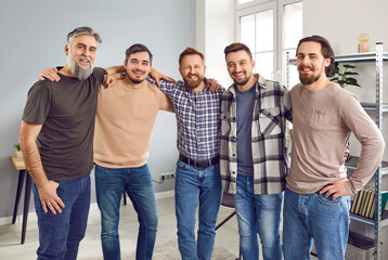 Smiling five male friends standing and hugging shoulders together. Portrait of positive cheerful hipster men wearing casual clothes smiling at camera indoors. Male friendship concept