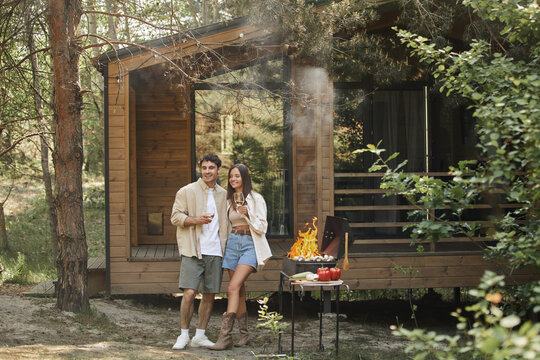 Positive romantic couple holding wine and standing near grill and vacation house at background