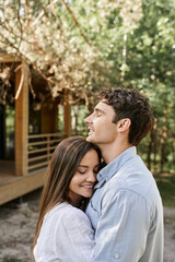 Obraz premium Smiling couple with closed eyes embracing and standing near vacation house at background outdoors