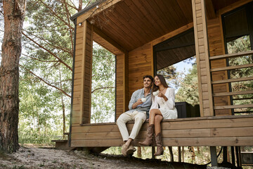 happy couple holding cups of coffee, man and woman smiling on porch of vacation house, hot beverage