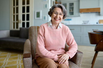 Happy smiling elderly woman sitting on chair in her own home livingroom