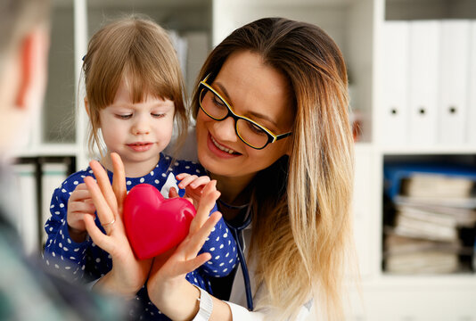 Female Doctor And Happy Little Child Hold In Arms Red Toy Heart Closeup. Cardio Therapeutist Student Education CPR 911 Life Save Physician Make Cardiac Physical Pulse Rate Measure Arrhythmia Lifestyle