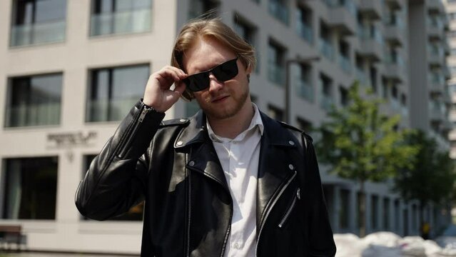 A fashionable man in sunglasses takes off his glasses and looks at the camera. A serious and brutal student. Slow motion, bottom angle. Architecture against the background of a man.